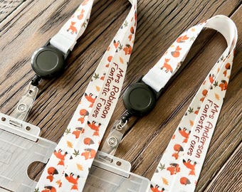 May include: Two white lanyards with a repeating pattern of orange foxes and autumn leaves. The lanyards have black retractable badge reels and clear plastic badge holders. Text on the lanyards reads "Mrs. Robinson Fantastic Foxes" and "Mrs. Anderson Fantastic Foxes."