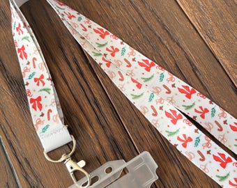 May include: A white lanyard with a festive Christmas pattern featuring red bows, green sprigs, and candy canes. The lanyard has a metal clasp and a clear plastic ID holder, perfect for holiday events or everyday use.