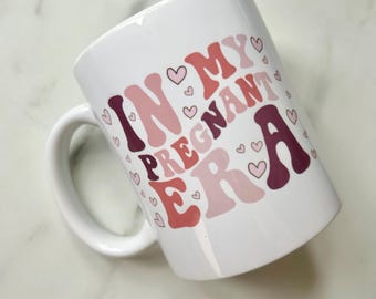 May include: White ceramic mug with the text "In My Pregnant Era" in pink, red, and brown lettering with pink hearts surrounding the text.