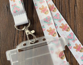 May include: A white lanyard with a repeating pattern of gingerbread people and wrapped gifts in pink and mint green. The lanyard has a silver-colored clasp and a clear plastic ID holder.
