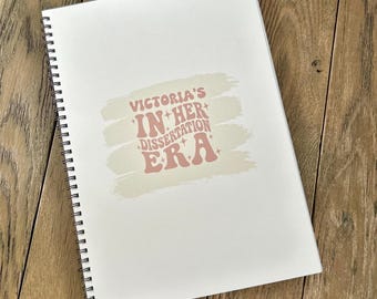 May include: A white spiral bound notebook with a pink and white design that says "Victoria's In Her Dissertation Era".