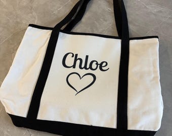 May include: White canvas tote bag with black trim and handles. The word "Chloe" is printed in black script above a black heart design. The bag is rectangular and appears to be made of a sturdy material.
