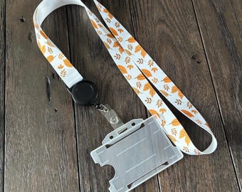 May include: A white lanyard with a fall leaf pattern in orange and yellow. The lanyard has a black retractable reel and a clear plastic ID holder. The lanyard is on a dark wood surface.