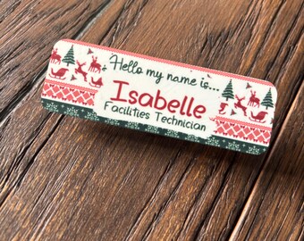 May include: A rectangular name tag with a festive Christmas design. The tag features the name "Isabelle" and the title "Facilities Technician." The design includes reindeer, Christmas trees, and a red and green pattern.