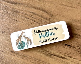 May include: A white rectangular name tag with a brown background. The tag features a cartoon illustration of two giraffes standing on a green and blue ball of yarn. The text on the tag reads "Hello my name is... Kaitlin Staff Nurse".