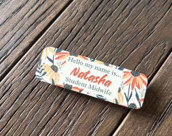 May include: A white name tag with a floral design featuring orange and yellow flowers. The text on the tag reads "Hello my name is... Natasha Student Midwife".
