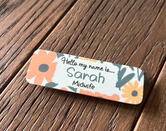 May include: A white name tag with a floral design in shades of orange, yellow, and green. The text on the tag reads "Hello my name is... Sarah Midwife".