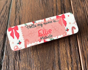 May include: A rectangular name tag with a festive design. The tag features the name "Ellie" and the title "Midwife" in red, with the phrase "Hello my name is..." above. The background is white with red bows, candy canes, and holly.