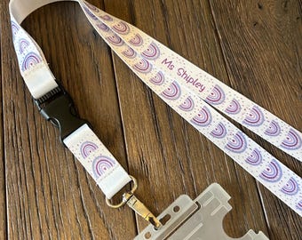 May include: White lanyard with a purple and white rainbow pattern and the text "Ms Shipely". The lanyard has a black plastic clip and a silver swivel hook. A clear plastic ID badge holder is attached to the lanyard.