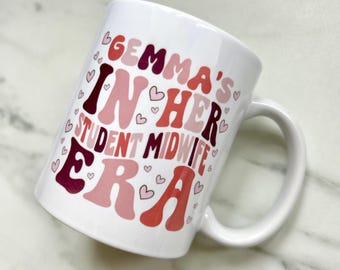 May include: White ceramic mug with the text "Gemma's In Her Student Midwife Era" in pink, red, and burgundy lettering with pink hearts surrounding the text.