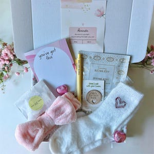 May include: A gift box containing self-care items. It features a pink bow headband, white fuzzy socks with a heart, a notepad with "you've got this", a pen, a face mask, and a small wooden token. The box also includes pink heart-shaped sweets.