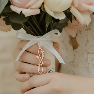 May include: A bouquet of pink and white roses and greenery, tied with a white ribbon. A gold-colored charm with the names "Sarah" and "Joshua" is attached to the ribbon. The bouquet is held by a person wearing a ring.