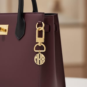 May include: A burgundy handbag with black handles and gold-colored hardware. A gold charm with the initials "MRB" hangs from the side. The bag has a structured shape and a rectangular design.