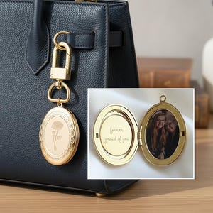 May include: A gold-colored oval locket keychain with a floral design and a photo inside. The locket is attached to a dark blue leather handbag. The locket is open, revealing a photo and the inscription "forever proud of you."