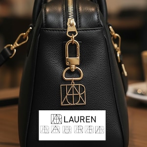 May include: Black leather handbag with gold-coloured hardware, including a zip and a decorative charm. The charm features the name "LAUREN" in a stylized font. The bag has a top handle and a shoulder strap.