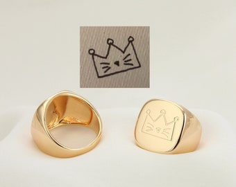 May include: Gold signet ring with a crown and cat face design engraved on the top. The ring is shown from two different angles.
