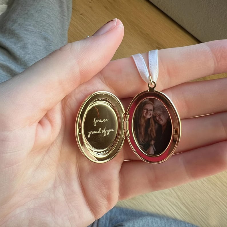 Może przedstawiać: Złoty owalny medalion z białą wstążką. Medalion jest otwarty, ukazując małe zdjęcie i wygrawerowane słowa "forever proud of you". Medalion jest trzymany w dłoni.