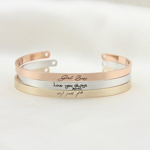 May include: Three stacked bracelets with different inscriptions. The top bracelet is rose gold and says "Girl Boss". The middle bracelet is silver and says "Love you always xoxo". The bottom bracelet is gold and has an inscription in a foreign language.