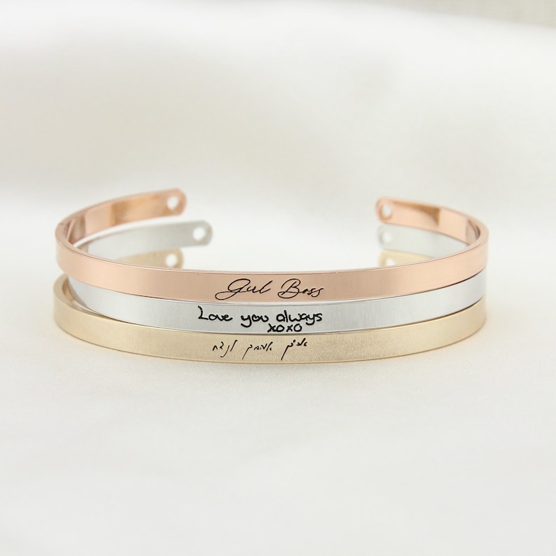 May include: Three stacked bracelets with different inscriptions. The top bracelet is rose gold and says "Girl Boss". The middle bracelet is silver and says "Love you always xoxo". The bottom bracelet is gold and has an inscription in a foreign language.