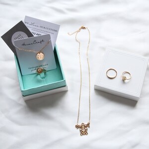 May include: A gold necklace with a turtle pendant, a gold ring with a heart-shaped stone, and a gold ring with a round stone. The jewelry is displayed on a white surface and in a turquoise box with a card that says "Horan Craft" and "Handmade with love".