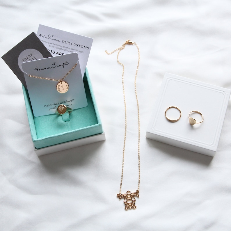 May include: A gold necklace with a turtle pendant, a gold ring with a heart-shaped stone, and a gold ring with a round stone. The jewelry is displayed on a white surface and in a turquoise box with a card that says "Horan Craft" and "Handmade with love".