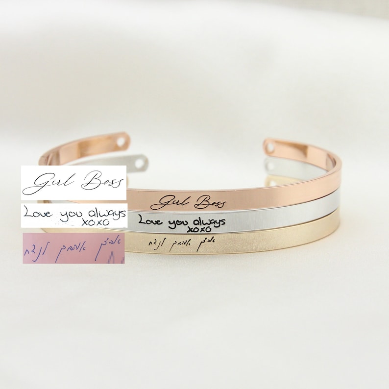 May include: Three stacked gold bracelets with the inscription "Girl Boss" on the top bracelet, "Love you always xoxo" on the middle bracelet, and Hebrew text on the bottom bracelet.