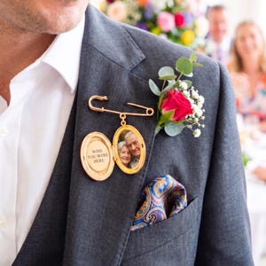 May include: A gold-colored locket with a photo of an elderly couple, attached to a gray suit jacket with a safety pin. The locket is open, revealing the photo and the text "WISH YOU WERE HERE". A red rose boutonniere and a patterned pocket square complete the look.