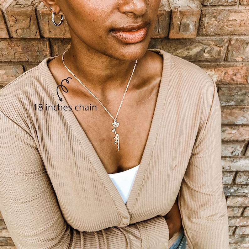 May include: A silver chain necklace with a delicate flower pendant. The chain is 18 inches long. The necklace is worn by a person with brown skin and a tan cardigan.