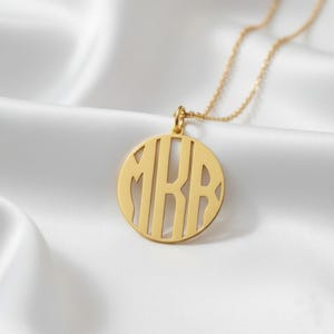 Puede incluir: Un collar con monograma dorado con un colgante circular. El colgante presenta las letras "MRB" recortadas en una fuente estilizada. El collar cuelga de una delicada cadena dorada, descansando sobre una superficie de satén blanco.