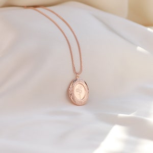 May include: A rose gold locket necklace with a delicate floral design. The locket is oval shaped and has a detailed border. The necklace is on a white fabric background.