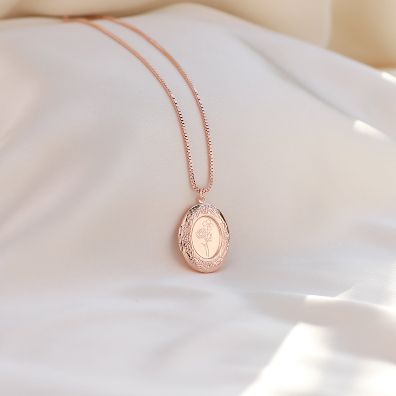 May include: A rose gold locket necklace with a delicate floral design. The locket is oval shaped and has a detailed border. The necklace is on a white fabric background.