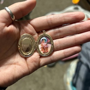 May include: Gold oval locket with a photo of a young child wearing a white hat and a pink dress inside.