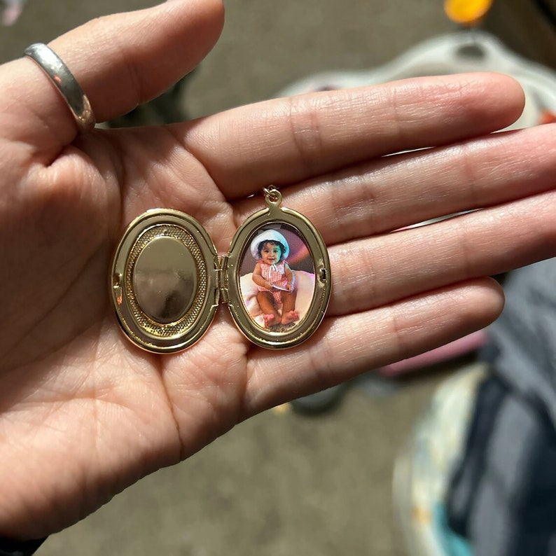 May include: Gold oval locket with a photo of a young child wearing a white hat and a pink dress inside.