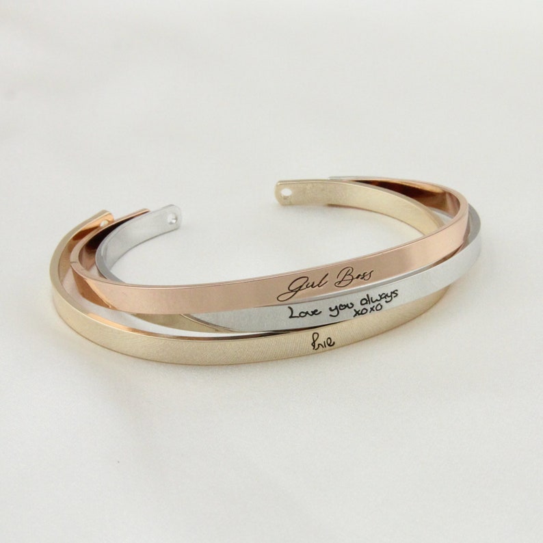May include: A gold, silver, and rose gold bangle bracelet with the inscription "Girl Boss" and "Love you always xoxo" in cursive handwriting.