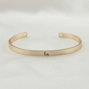 May include: A gold bangle bracelet with the word "brie" inscribed on it.