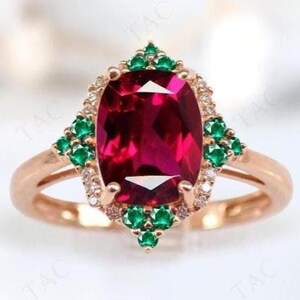 Puede incluir: Un anillo de oro rosa con una gran piedra de rubí de forma ovalada rodeada por un halo de diamantes más pequeños y esmeraldas verdes.