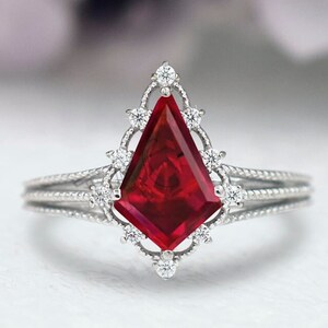 Puede incluir: Un anillo de plata con una gran piedra preciosa roja facetada engastada en un diseño delicado y ornamentado. El anillo está rodeado de piedras preciosas más pequeñas y transparentes.