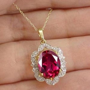 May include: A gold pendant necklace featuring an oval, deep red gemstone. The gemstone is surrounded by a halo of small, clear stones set in a decorative, openwork frame. The necklace has a delicate gold chain.
