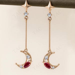 Peut inclure: Paire de boucles d'oreilles pendantes dorées. Chaque boucle d'oreille présente un sommet en forme d'étoile, une pierre précieuse transparente, une longue barre verticale, un croissant de lune avec des pierres précieuses transparentes et une pierre précieuse rouge en forme de goutte.