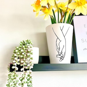 May include: White ceramic vase with a black line drawing of two hands holding each other. The vase is filled with yellow daffodils and is sitting on a black shelf.