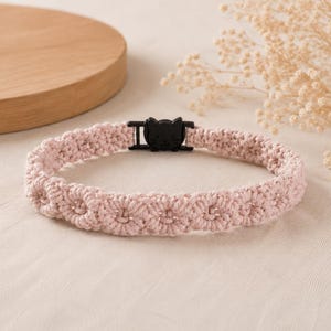 May include: A light pink crocheted collar featuring a repeating floral pattern throughout the band. The collar is secured with a black plastic buckle shaped like a cat head, providing a functional closure for the woven textile accessory.