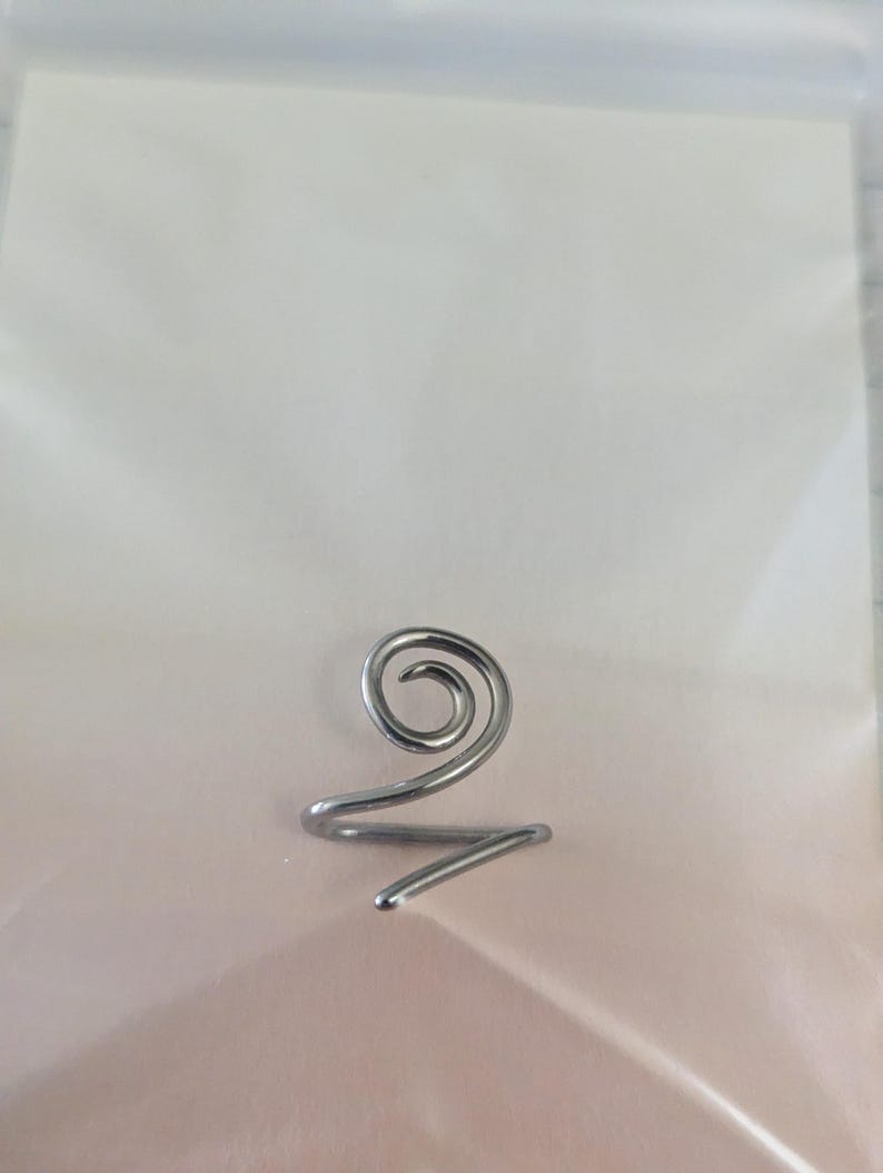 K&ouml;nnte beinhalten: Ein Ring aus silberfarbenem Metall, der aus einem einzigen durchgehenden Draht in Spiralform besteht. Der Draht biegt sich nach innen zu einer kreisf&ouml;rmigen Windung an der Oberseite, w&auml;hrend das restliche Ende eine einfache, offene Ringschiene bildet.