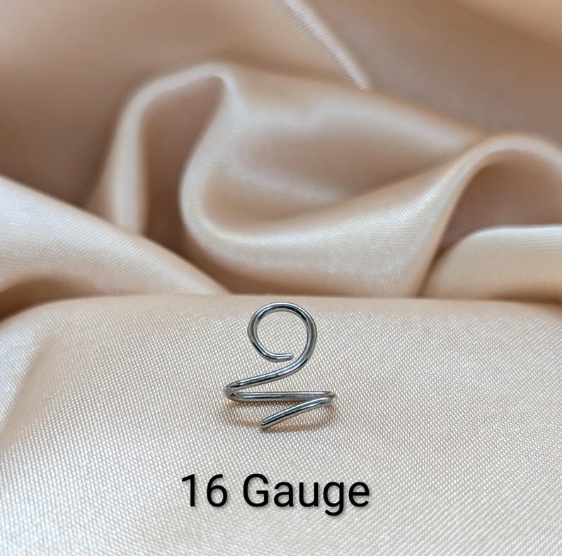 K&ouml;nnte beinhalten: Ein silberfarbener Ohrring aus Metall mit einem Spiraldesign, das oben eine einzelne Schlaufe und ein spitz zulaufendes Ende aufweist. Das St&uuml;ck ist deutlich mit dem Text 16 Gauge beschriftet, was die Dicke angibt, und besteht aus glattem Draht.