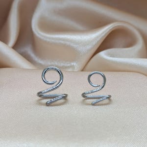 K&ouml;nnte beinhalten: Zwei silberfarbene Ohrklemmen aus Metall, die jeweils ein einfaches Spiraldesign aufweisen. Die Klemmen bestehen aus einem durchgehenden Draht, der sich oben zu einer kreisf&ouml;rmigen Schlaufe biegt und unten in einem geraden Stab endet.