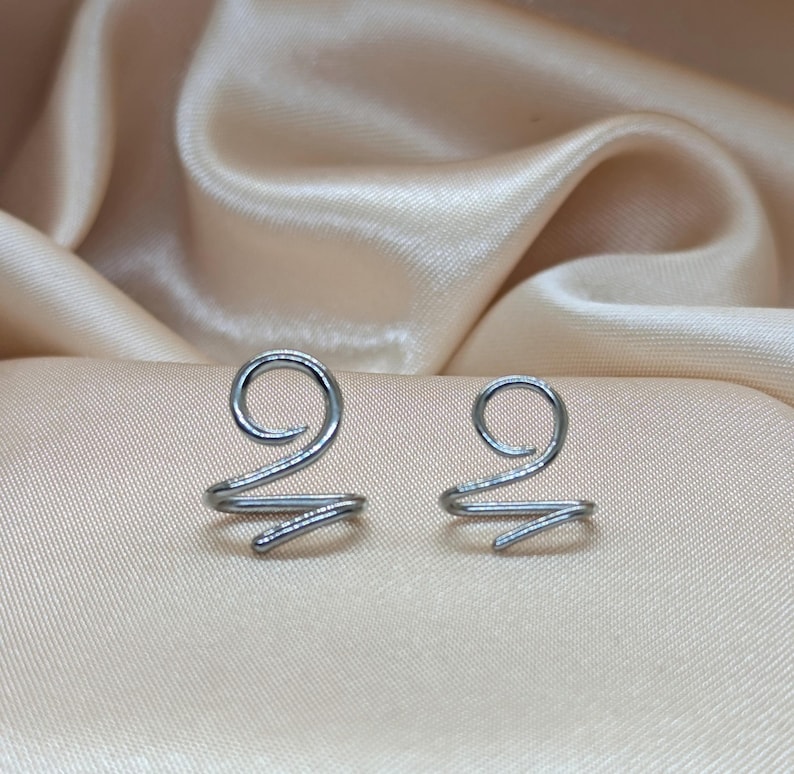 K&ouml;nnte beinhalten: Zwei silberfarbene Ohrklemmen aus Metall, die jeweils ein einfaches Spiraldesign aufweisen. Die Klemmen bestehen aus einem durchgehenden Draht, der sich oben zu einer kreisf&ouml;rmigen Schlaufe biegt und unten in einem geraden Stab endet.