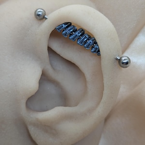 K&ouml;nnte beinhalten: Ein industrielles Ohrpiercing mit einem Mittelst&uuml;ck, das mit blauen Metalldrahtwicklungen verziert ist. Das Piercing wird an beiden Enden durch glatte, polierte silberfarbene Kugeln fixiert und verl&auml;uft durch den oberen Knorpel des Ohrs.