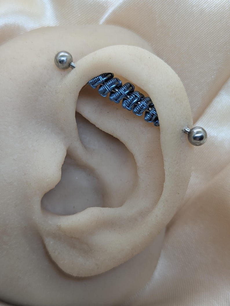K&ouml;nnte beinhalten: Ein industrielles Ohrpiercing mit einem Mittelst&uuml;ck, das mit blauen Metalldrahtwicklungen verziert ist. Das Piercing wird an beiden Enden durch glatte, polierte silberfarbene Kugeln fixiert und verl&auml;uft durch den oberen Knorpel des Ohrs.