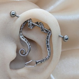K&ouml;nnte beinhalten: Ein silberfarbenes Industrial-Barbell-Piercing bestehend aus einem langen, geraden Stab mit kugelf&ouml;rmigen Enden und einem angebrachten, aufwendig drahtgewickelten Dekorationselement, das sich spiralf&ouml;rmig im Ohrknorpel nach unten windet.
