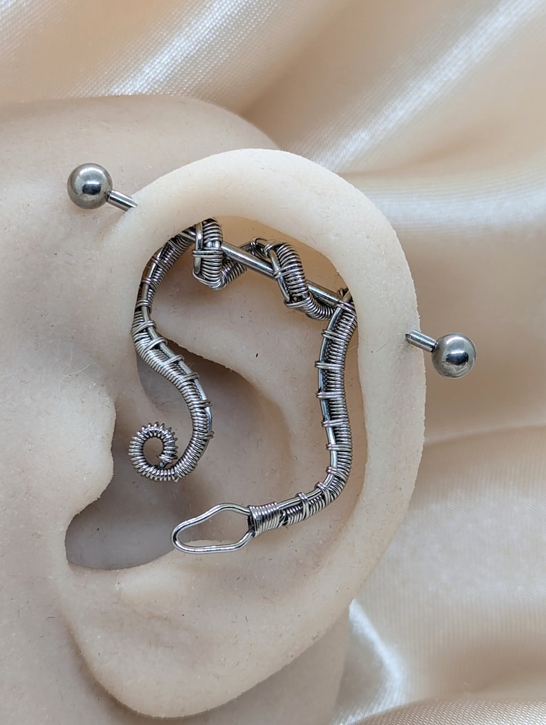 K&ouml;nnte beinhalten: Ein silberfarbenes Industrial-Barbell-Piercing bestehend aus einem langen, geraden Stab mit kugelf&ouml;rmigen Enden und einem angebrachten, aufwendig drahtgewickelten Dekorationselement, das sich spiralf&ouml;rmig im Ohrknorpel nach unten windet.