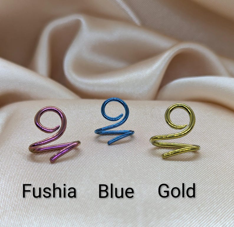 K&ouml;nnte beinhalten: Drei spiralf&ouml;rmige Haarspangen aus Draht in Fuchsia, Blau und Gold. Jedes St&uuml;ck hat ein gewundenes Design mit einer Schlaufe an der Oberseite und einem spitz zulaufenden Ende. Der Text Fushia, Blue und Gold steht unter dem jeweiligen Haarschmuck.